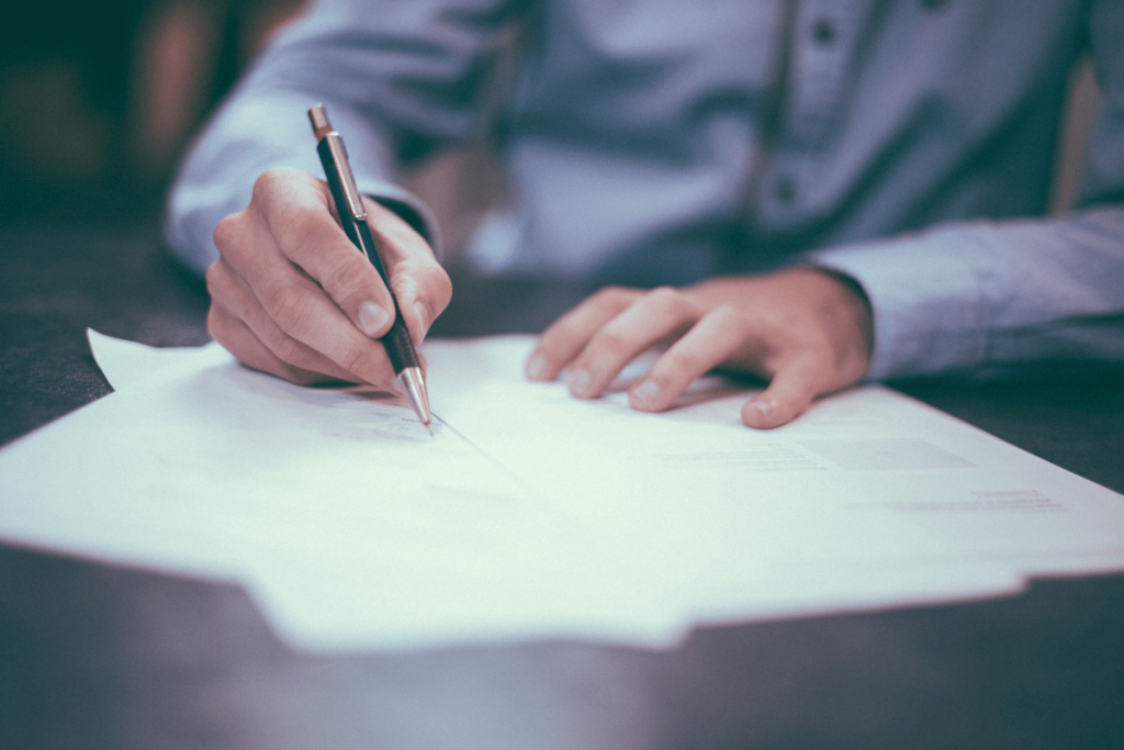 Person signing a document with a pen on a wooden table with a blurred background.