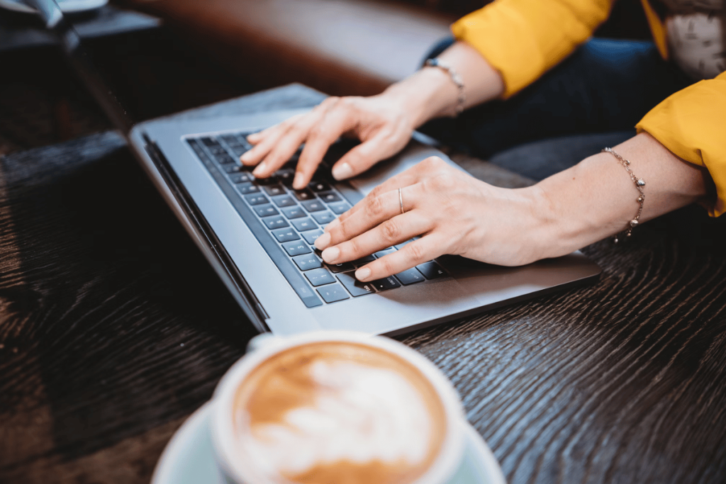 Close-up of hands typing on a laptop keyboard next to a cappuccino.